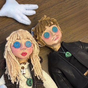 May include: Two handmade dolls with yarn hair and blue button eyes. The left doll has blonde hair, a white textured top, and a brown vest. The right doll has brown hair, a black jacket, and a green pendant necklace. A white plush hand is visible.