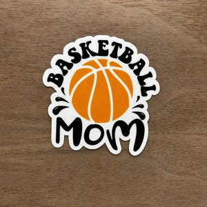 May include: White sticker with black text that reads "Basketball Mom" with a basketball graphic in the center.