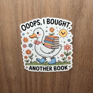 May include: A sticker featuring a cartoon duck wearing blue boots and a pink bow tie, carrying a stack of books. The sticker has the text "OOOPS, I BOUGHT ANOTHER BOOK" with flowers and a smiling sun.