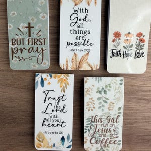 May include: A collection of rectangular bookmarks with inspirational quotes and floral designs. One bookmark reads "But first pray," another says "With God, all things are possible," and a third says "Faith Hope Love."