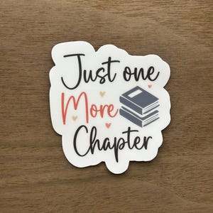 May include: White sticker with black text that reads "Just one more chapter" with a stack of three books in the middle of the text. The books are gray with a black spine. There are small red hearts around the text and books.