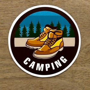May include: A round sticker with a black background and white outline. The sticker features a pair of brown hiking boots with yellow laces and a blue and green forest scene in the background. The text "CAMPING" is written in white letters below the boots.
