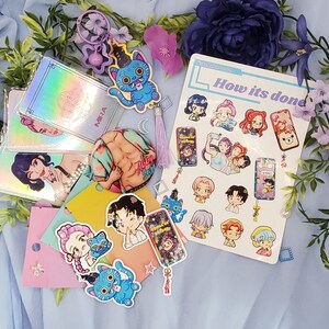 May include: A collection of stickers, keychains, and holographic cards featuring anime-style illustrations. The items include images of cartoon characters, cats, and the text "How its done". The items are arranged on a blue fabric background with floral accents.