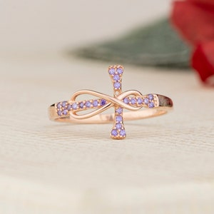 May include: A rose gold ring with a cross design, featuring a purple infinity symbol and small purple gemstones.