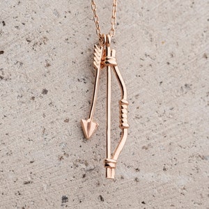 May include: A gold necklace with a bow and arrow pendant. The pendant is made of metal and has a detailed design.