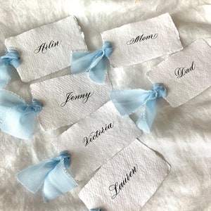 Marque-places blancs ou ivoire avec rubans de soie bleu layette, porte-noms à bords bruts sur papier de coton fait main