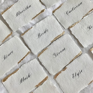 May include: White place cards with gold accents and black calligraphy. The names on the cards include:  Marisha, Bride, Groom, Bruce, Mark, Mel, Genevieve, Caitriona, and Elyse.