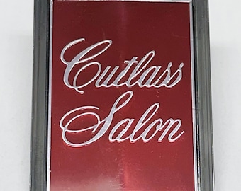 May include: A rectangular emblem with a shiny silver frame and a deep red center. The words "Cutlass Salon" are written in a cursive, silver font. The emblem is likely a car badge.