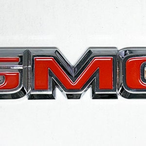Könnte beinhalten: Ein verchromtes und rotes GMC-Emblem. Das Emblem zeigt die Buchstaben "GMC" in einer kräftigen, stilisierten Schriftart. Die Buchstaben sind in Chrom umrandet, mit roter Innenseite. Das Emblem ist rechteckig und hat eine glänzende Oberfläche.
