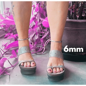 May include: A pair of silver anklets with a 6mm width, worn on a person's ankles. The anklets are simple and elegant, with a smooth, polished finish.