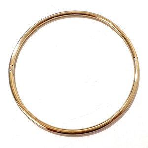 K&ouml;nnte beinhalten: Eine goldfarbene, kreisf&ouml;rmige Halskette mit einem Verschluss. Die Halskette hat ein schlichtes, elegantes Design mit einer glatten, polierten Oberfl&auml;che. Der Schmuck ist vor einem wei&szlig;en Hintergrund platziert.