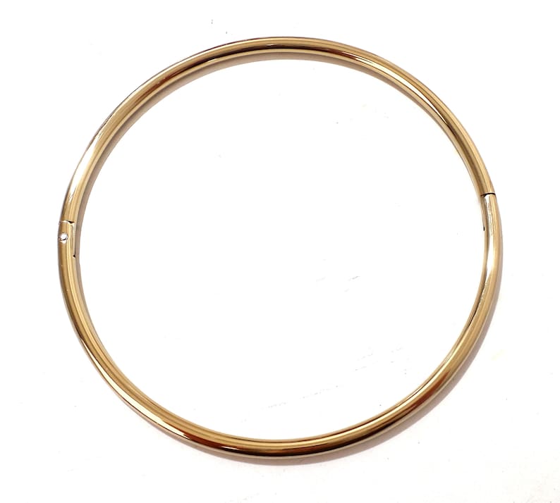 K&ouml;nnte beinhalten: Eine goldfarbene, kreisf&ouml;rmige Halskette mit einem Verschluss. Die Halskette hat ein schlichtes, elegantes Design mit einer glatten, polierten Oberfl&auml;che. Der Schmuck ist vor einem wei&szlig;en Hintergrund platziert.