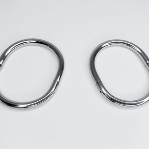 Op de afbeelding: Twee zilveren metalen ringen, elk met een gladde, afgeronde ovale vorm. De ringen zijn open en lijken uit één stuk metaal te zijn gemaakt.