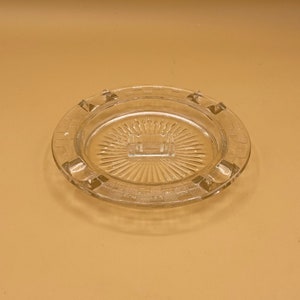 May include: A clear glass ashtray with a decorative, sunburst pattern in the center and four indentations around the rim.