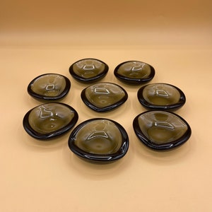 May include: Set of eight small, clear glass dishes with a dark brown rim. The dishes are arranged in a circle on a light brown surface.