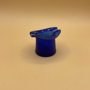May include: A small, blue glass ashtray shaped like a top hat.