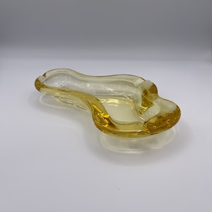 May include: A yellow glass ashtray with a wavy, organic shape. The ashtray is designed to hold cigarettes and is made of clear glass with a yellow tint.