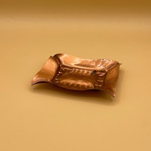 May include: A small, copper-colored tray with a wavy, hammered design. The tray has a slightly raised edge and a decorative pattern around the center.