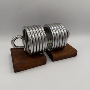 May include: Two stainless steel mugs with looped handles, each resting on a small, dark wooden base. The mugs are made of coiled metal, giving them a unique, industrial look. The background is plain white.