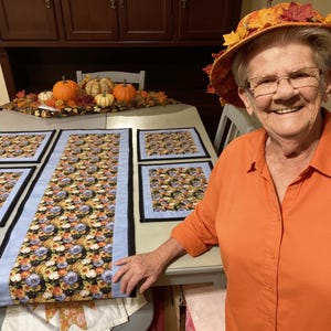 Op de afbeelding: Een tafelloper en vier bijpassende placemats met een zwarte stoffen basis en een kleurrijk patroon van pompoenen, zonnebloemen en herfstbladeren. Elk stuk is afgewerkt met een lichtblauwe stoffen rand en een dunne zwarte bies.