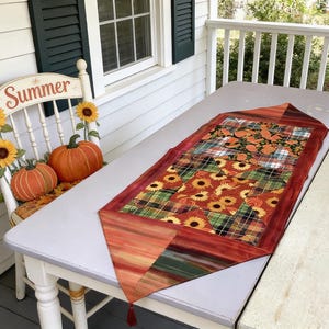 Puede incluir: Camino de mesa con temática otoñal con un diseño de patchwork con girasoles y cuadros. El camino de mesa está colocado sobre una mesa blanca. Una silla blanca con la palabra "Summer" en el respaldo y girasoles y calabazas decorativas en el fondo.