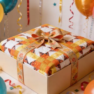 May include: A folded quilt featuring a geometric star pattern in shades of yellow, orange, and white, placed inside a square cardboard box. The quilt is wrapped with a decorative tan ribbon featuring a floral print and topped with a large bow.