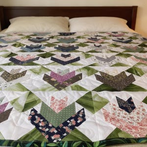 May include: A handmade quilt with a floral pattern. The quilt features a white background with green, pink, blue, and brown tulip-shaped designs. The quilt is on a bed with white pillows and a dark brown headboard. The quilt's dimensions are not visible.