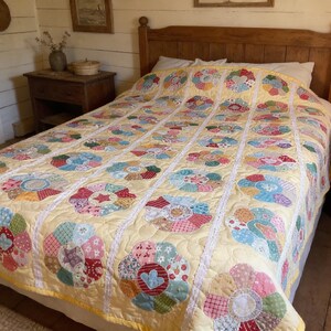 May include: A handmade quilt with a yellow background and colorful floral appliqués. The quilt features a repeating pattern of large, circular flower designs in shades of red, blue, pink, and green, with white lace trim. The quilt is on a wooden bed frame.