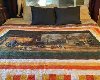 May include: A quilted bedspread featuring a rural scene with a tractor, a small wagon, and a truck. The quilt has a border of horizontal stripes in orange, yellow, and white. Two dark blue pillows rest on the bed.