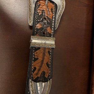 May include: A brown leather belt with a silver buckle and a silver tip. The belt has a leaf pattern embossed into the leather.
