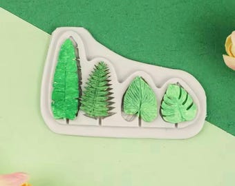Puede incluir: Molde de silicona con cinco formas de hojas, incluyendo un helecho, una hoja de palma y tres tipos diferentes de hojas. El molde es blanco y las hojas son verdes.