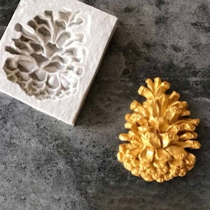 Op de afbeelding: Een witte siliconen mal voor het maken van een cakedecoratie in de vorm van een dennenappel. De mal wordt getoond naast een gouden dennenappel cakedecoratie.