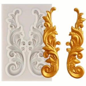 以下が含まれることがあります： 2つの装飾的なケーキトッパーを作るためのシリコンモールド。モールドは白で、詳細で装飾的なデザインが施されています。モールドは、モールドを使用して作られた2つの金色のケーキトッパーと共に示されています。