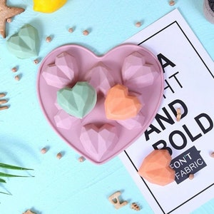 May include: A heart-shaped silicone mold filled with geometric heart-shaped candies in shades of pink, green, and orange. The mold sits on a light blue surface with a lemon and a sign that reads "A HT AND BOLD".
