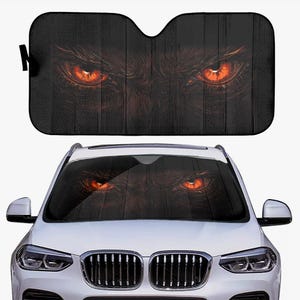May include: Black car windshield sunshade featuring a close-up of glowing orange eyes. The eyes are set against a dark background, creating a striking and intimidating design. The sunshade is designed to fit a car windshield.