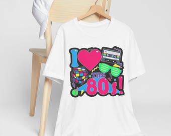 May include: White t-shirt with a colorful graphic that says "I love the 80's!" The design includes a pink heart, cassette tape, Rubik's cube, and neon green sunglasses. The shirt is hanging on a wooden hanger.