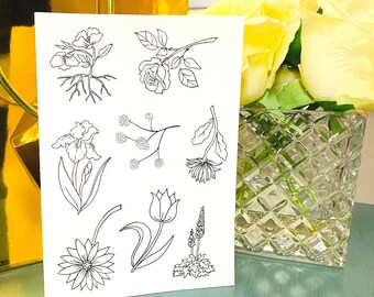 May include: A white card with black line drawings of flowers, such as a rose, iris, and tulip. The card is displayed, with a gold gift box and a vase of yellow roses in the background.
