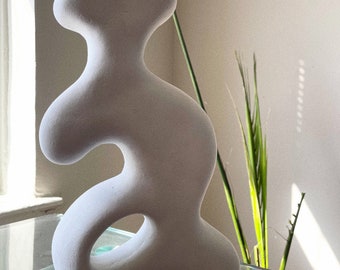 May include: A white ceramic sculpture with an abstract, organic shape. The sculpture has a hole in the middle and is resting on a glass surface.