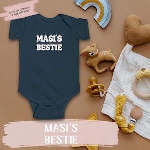 Peut inclure: Body bébé bleu marine avec l'inscription blanche "MASI'S BESTIE". L'image montre également une sucette, un hochet en bois, un cœur en peluche, un jouet en forme de cerf et une décoration en forme d'arc-en-ciel. Le texte "8 OPTIONS DE COULEUR 5 OPTIONS DE TAILLE" est en haut à gauche.