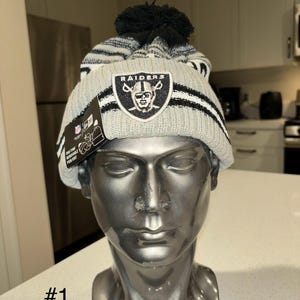 May include: A grey and black knitted beanie with a pom pom on top. The beanie has a black and white striped pattern and a Raiders logo on the front. The logo is a black and white image of a football player's head wearing a helmet. The beanie is on a mannequin's head.