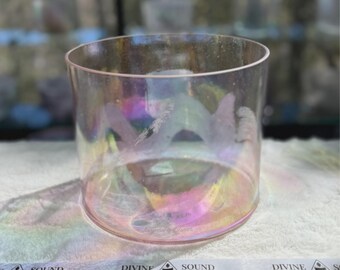 May include: A clear, iridescent glass singing bowl with a cylindrical shape. The bowl has a wide opening and a smooth, polished surface. The glass reflects a rainbow of colors. A white ribbon with the words "DIVINE SOUND" is in the foreground.