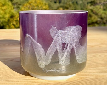 May include: A purple and silver crystal singing bowl with a white base. The bowl has a gradient effect, transitioning from purple to silver. The bowl has a silver abstract design and the text "Crystal Oasis".