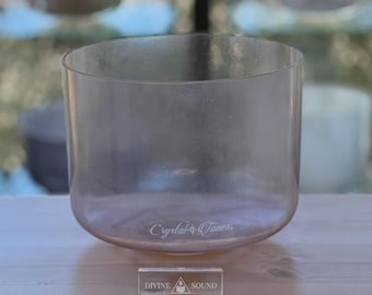 May include: A clear, cylindrical crystal singing bowl with rounded edges. The words "Crystal Tones" are etched on the bowl's base. A small rectangular sign below reads "DIVINE SOUND". The bowl sits on a wooden surface.