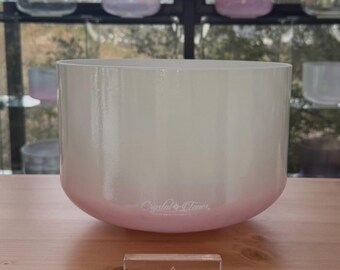 May include: A large, pearlescent crystal singing bowl with a soft pink ombre effect at the base. The bowl sits on a wooden surface, with a small rectangular block that reads "DIVINE SOUND". The bowl has the text "Crystal Tones" on the side.