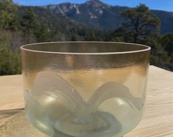 May include: A clear glass singing bowl with a gold rim and a frosted wave design. The bowl has a light blue tint at the bottom and the text "Crystal Tones" is visible. The bowl is sitting on a wooden surface.