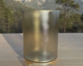 May include: A frosted gold-colored cylindrical glass candle holder. The candle holder is sitting on a wooden surface. A small, rectangular clear acrylic sign with the words "DIVINE SOUND" is in front of the candle holder.