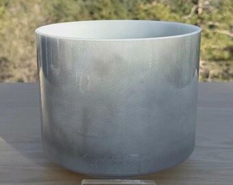 May include: A large, cylindrical, frosted glass singing bowl with a white interior. The bowl has a gradient effect, transitioning from a light gray at the top to a darker gray at the base. The words "Crystal Tones" are faintly visible. A clear rectangular block with the words "DIVINE SOUND" is in front.