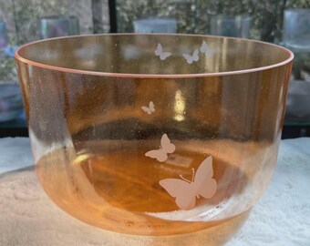May include: A large, translucent, peach-colored bowl with etched butterfly designs. The bowl has a wide, open shape and is likely made of glass or a similar material. The words "DIVINE SOUND" are printed on a white ribbon.