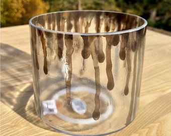 May include: A clear glass votive candle holder with brown dripped paint effect. The cylindrical candle holder has a thick base and is sitting on a wooden surface. The brown paint drips down the sides of the glass.