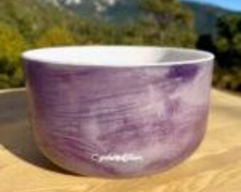 May include: A purple crystal singing bowl with a white interior. The bowl has a smooth, rounded shape and a marbled appearance. The text "Crystal Tones" is printed on the front of the bowl. The background features a blurred view of trees and mountains.